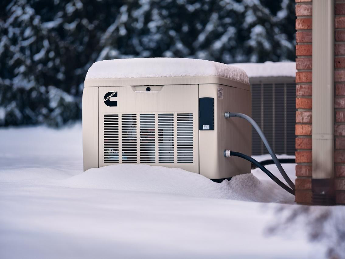 kenai-winter-backup-generator A Cummins home standby generator installed outside a brick home, partially covered in deep snow during an Alaskan winter.