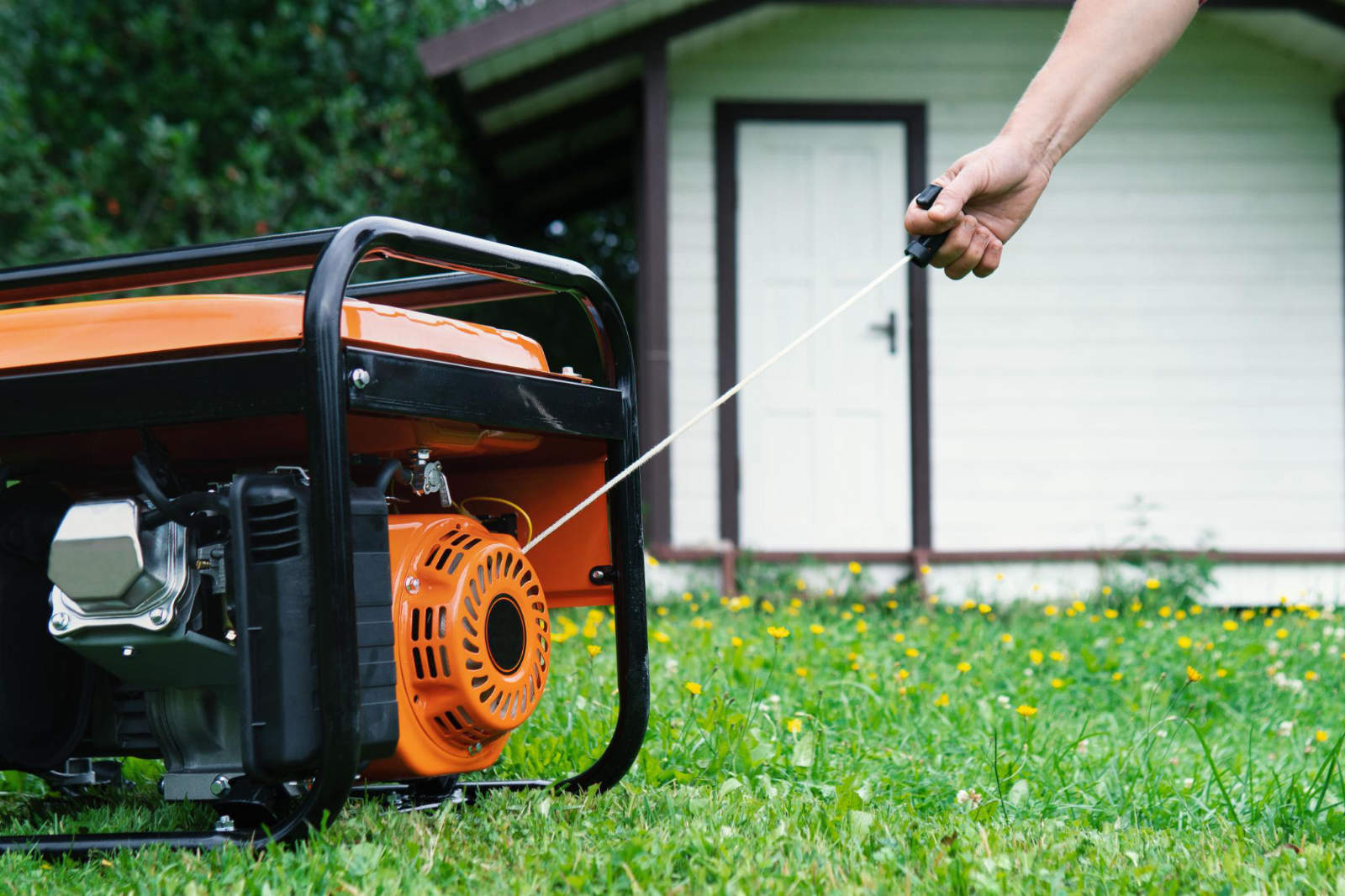 A person pulling the recoil starter cord on a portable orange generator sitting on a green lawn in front of a shed.