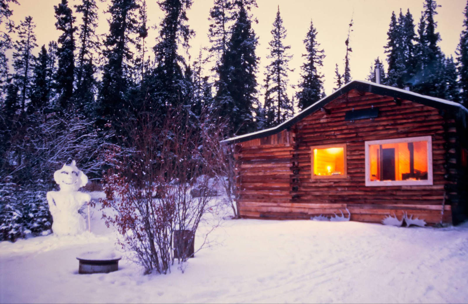 Warmly lit wooden log cabin in a snowy forest during winter in Alaska.
